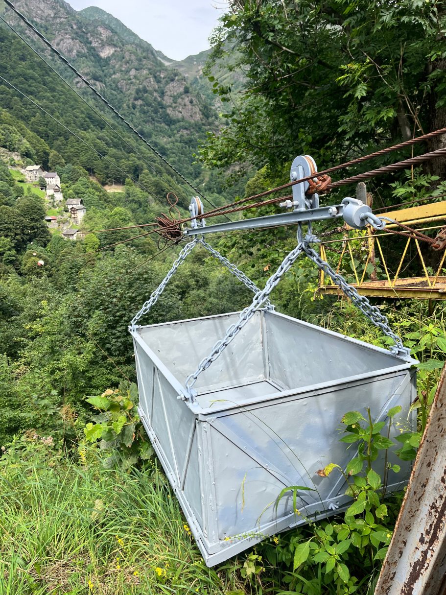 Eine metallene Seilbahnkabine schwebt über einer grünen Tallandschaft.