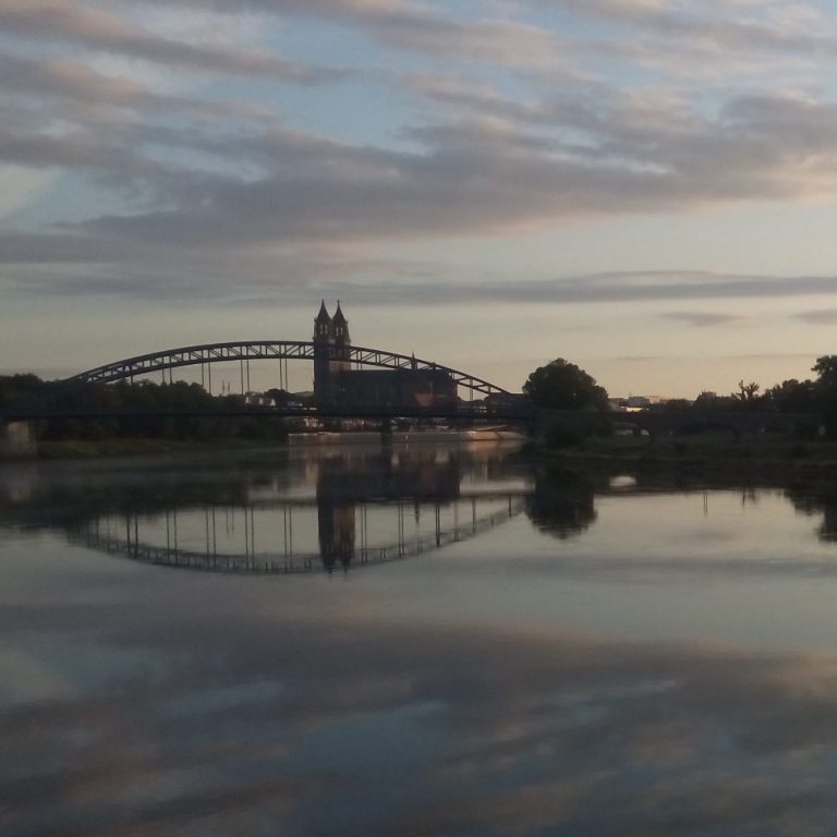 Brücke mit Türmen am Ufer eines ruhigen Gewässers bei Sonnenuntergang.