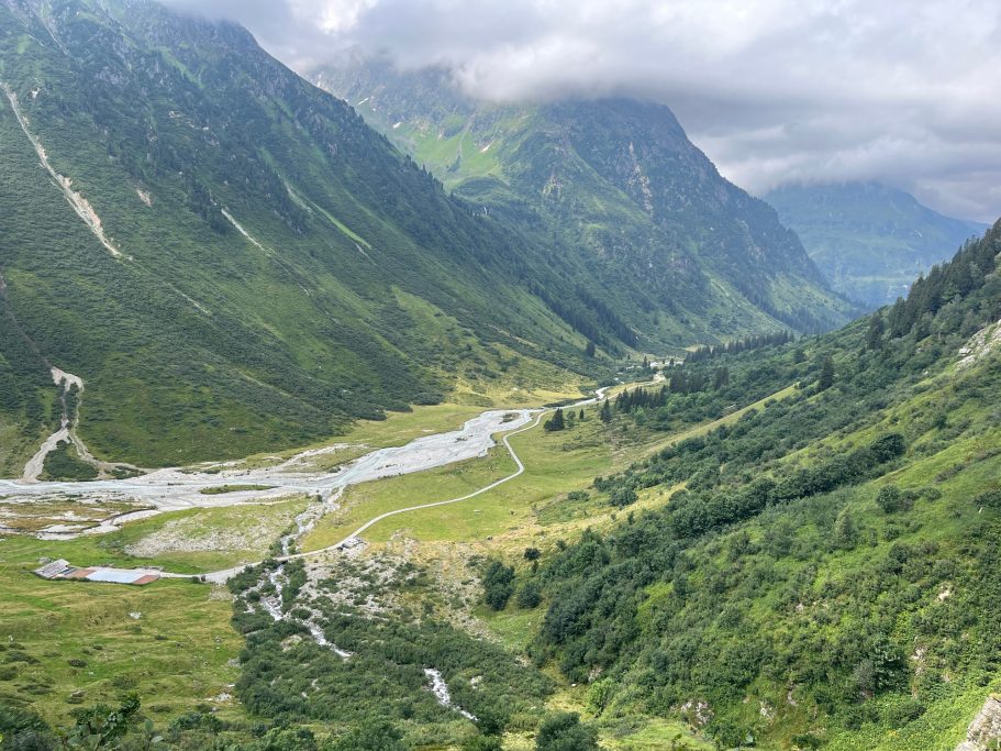 Mässboden Blick auf ein grünes Tal mit Fluss, umgeben von hohen Bergen und bewölktem Himmel.