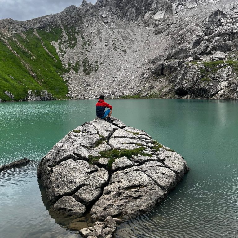 Person in rotem Pullover sitzt auf einem großen Stein am Ufer eines klaren Sees, umgeben von Bergen.