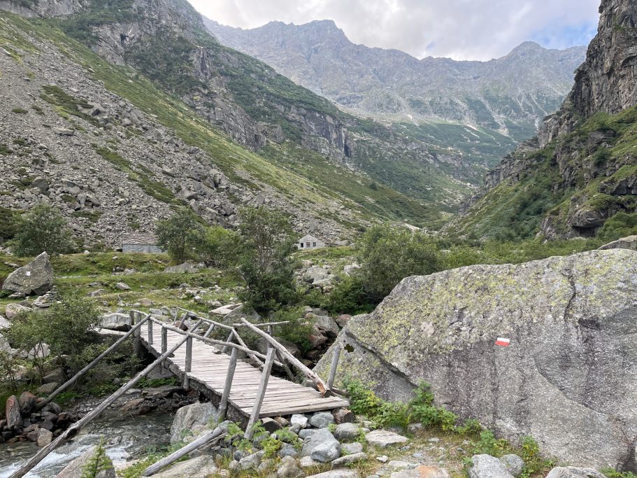 Val Vogna Eine Holzbrücke über einen schmalen Fluss, umgeben von Bergen und grüner Vegetation.