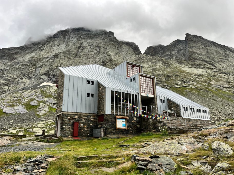 Rifugion Valanta Moderne Berghütte aus Metall und Stein, umgeben von hohen Bergen und wolkenverhangenem Himmel.