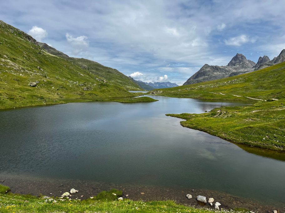Idyllische Landschaft mit einem ruhigen See, umgeben von grünen Hügeln und Bergen.