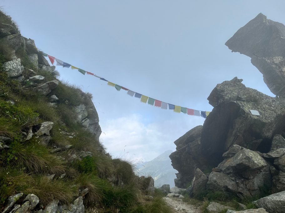 Bunte Gebetsfahnen hängen zwischen großen Felsen in einer nebligen Landschaft.