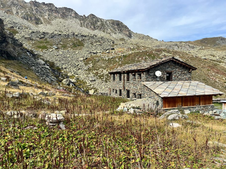Rifugio Alpetto Almhütte umgeben von Felsen und hohem Gras in montaner Landschaft.