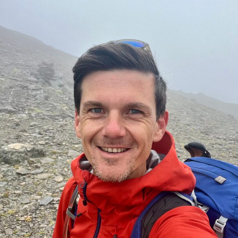 Ein lächelnder Mann in roter Jacke vor einer nebligen Berglandschaft.