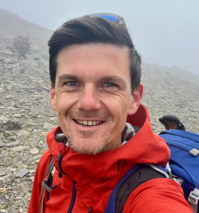 Mann mit roter Jacke und Rucksack, lächelt in einer bergigen, nebligen Landschaft.