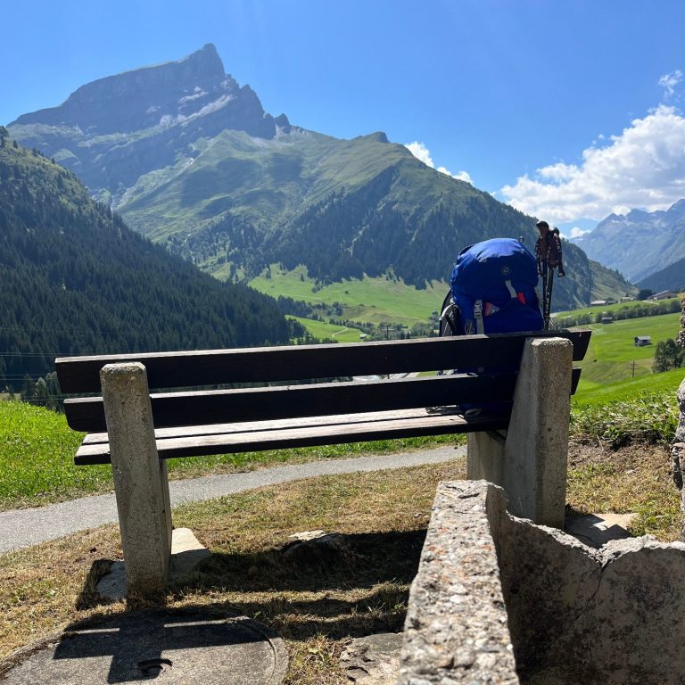Blick auf eine Bank in den Alpen mit grünen Wiesen und Bergen im Hintergrund.